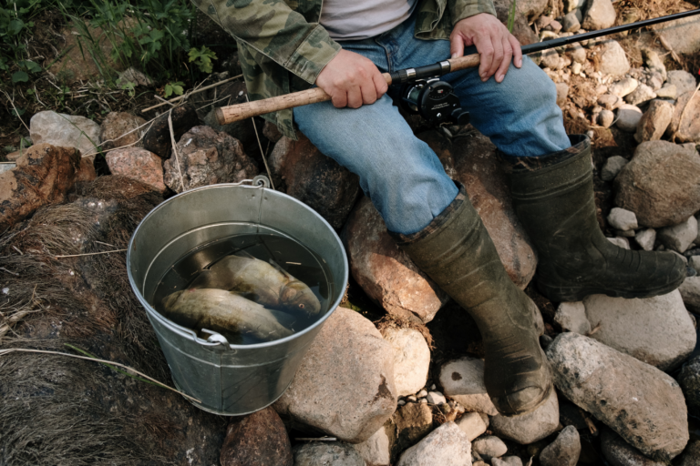 Eine Person sitzt an einem steinernen Untergrund und hält eine Angelrute, während ein Eimer mit Wasser und 2 Fischen daneben steht.